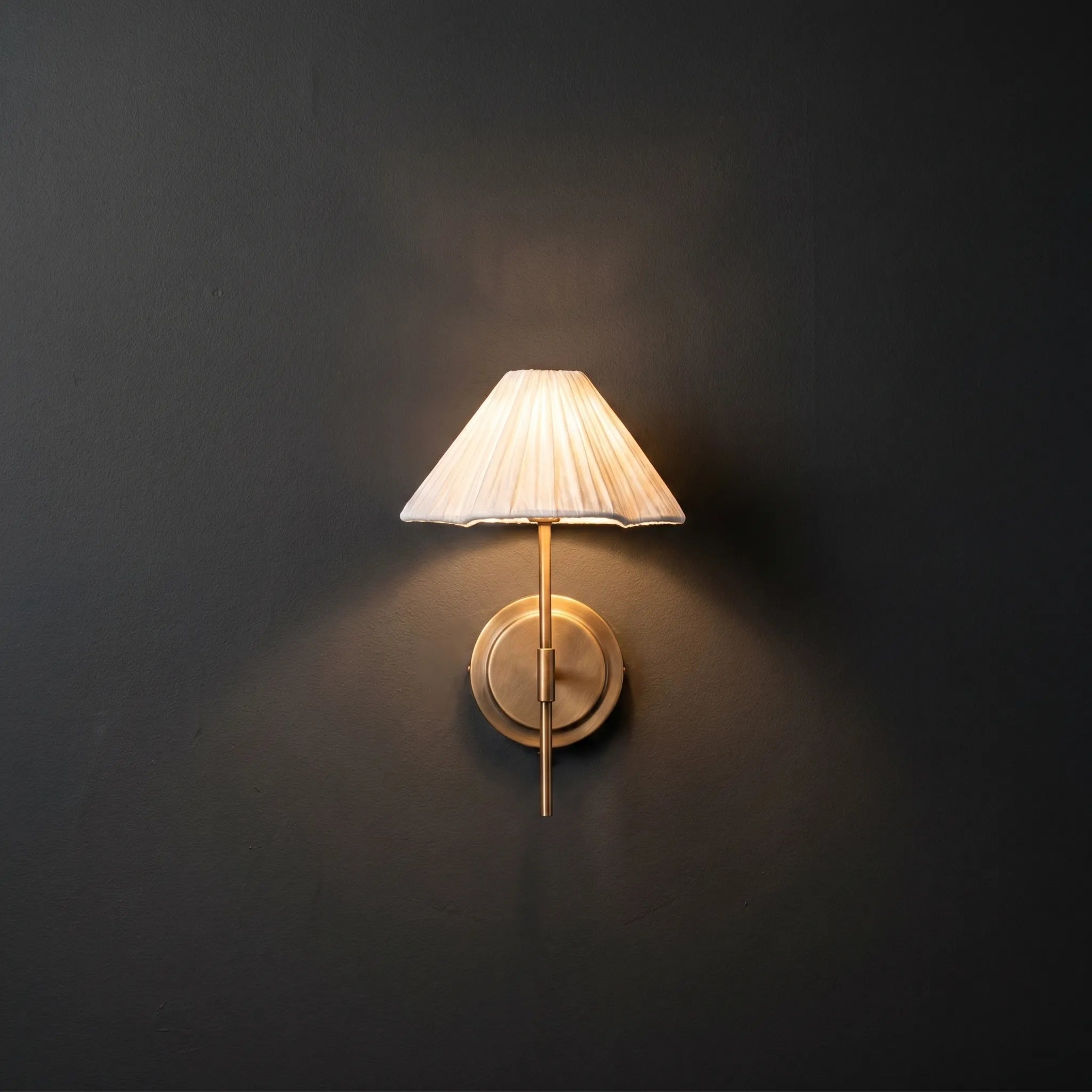 Pleated Fabric Wall Sconce – Brass Arm Wall Light with White Gathered Shade in a dark studio.
