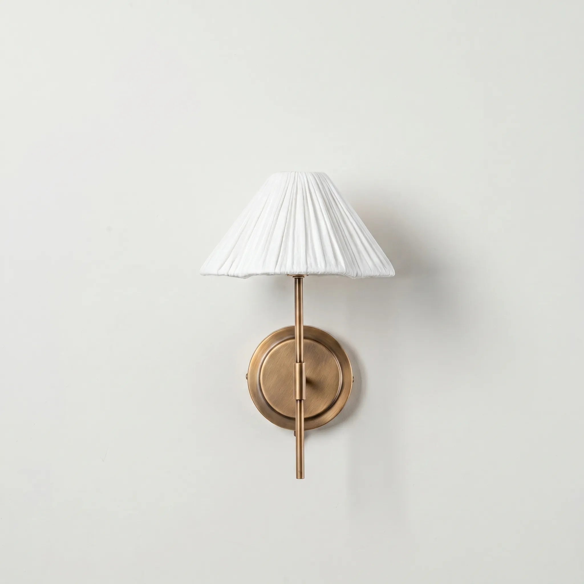 Pleated Fabric Wall Sconce – Brass Arm Wall Light with White Gathered Shade in a white studio. 