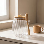 Clear glass jar with wooden lid and spoon on a light surface near a window.