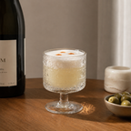 Cocktail in a textured glass with a bottle and bowl of olives on a wooden surface