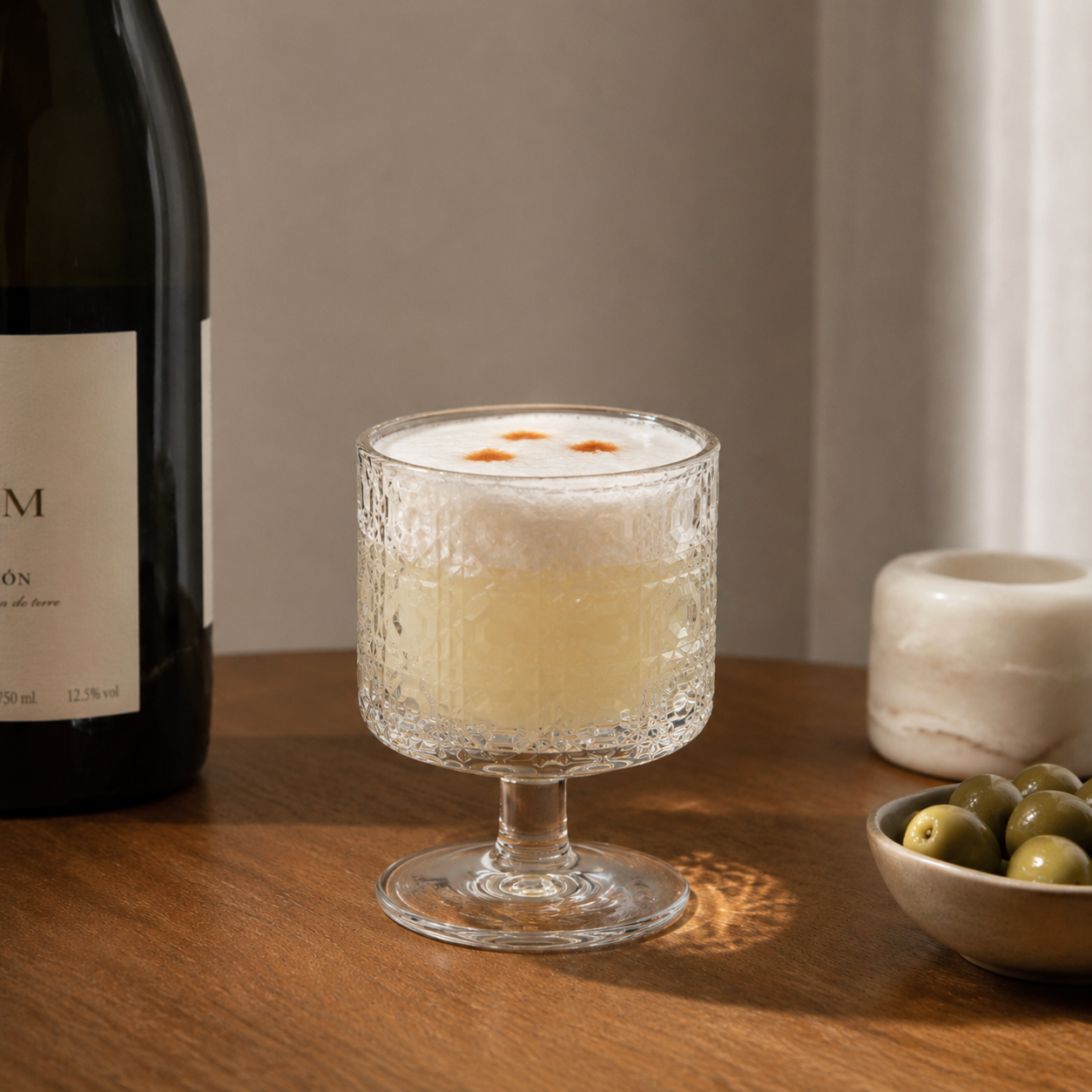 Cocktail in a textured glass with a bottle and bowl of olives on a wooden surface