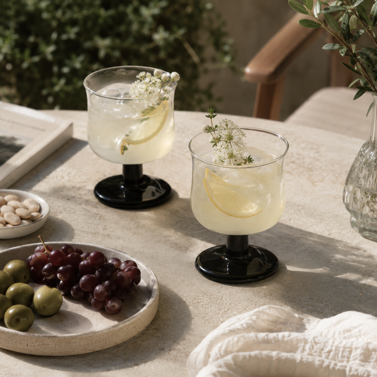 Two glasses of lemonade with ice and garnishes on a table with grapes and olives.