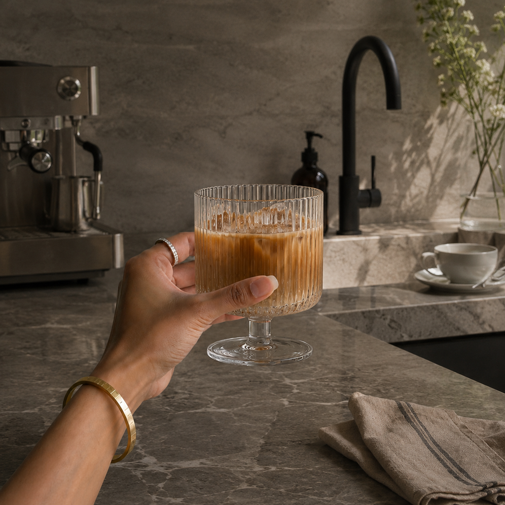 Hand holding a glass of iced coffee in a kitchen setting with a coffee machine and sink.
