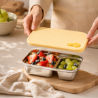 Person opening a yellow lid on a stainless steel bento box filled with fruits on a wooden cutting board.