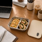 Lunchbox with compartments on a wooden desk with a laptop, books, and drinks.