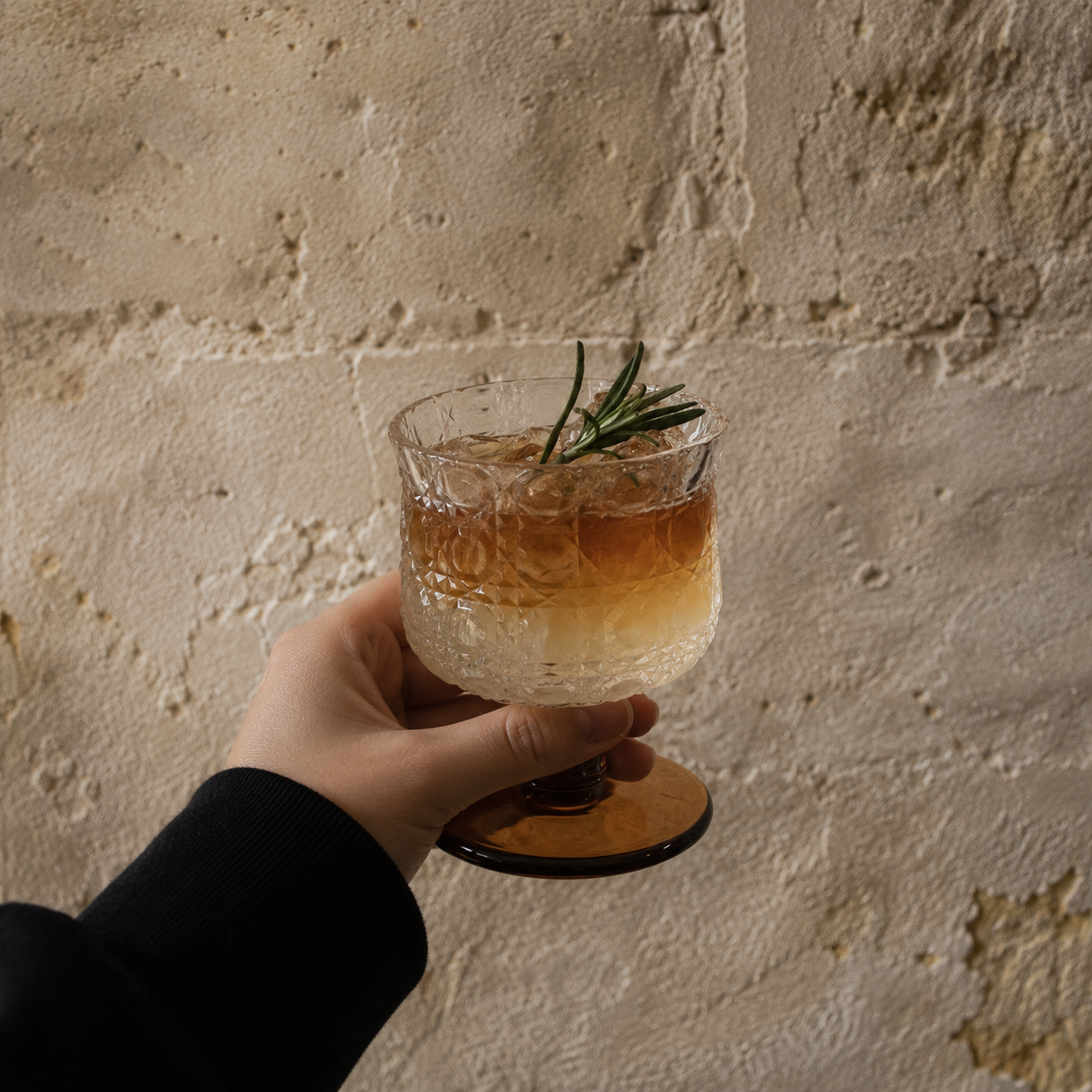 Hand holding a cocktail glass with a drink and a rosemary sprig against a textured stone wall.