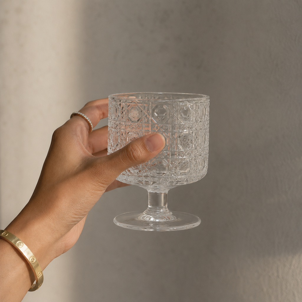 Hand holding a clear glass with intricate patterns against a neutral background