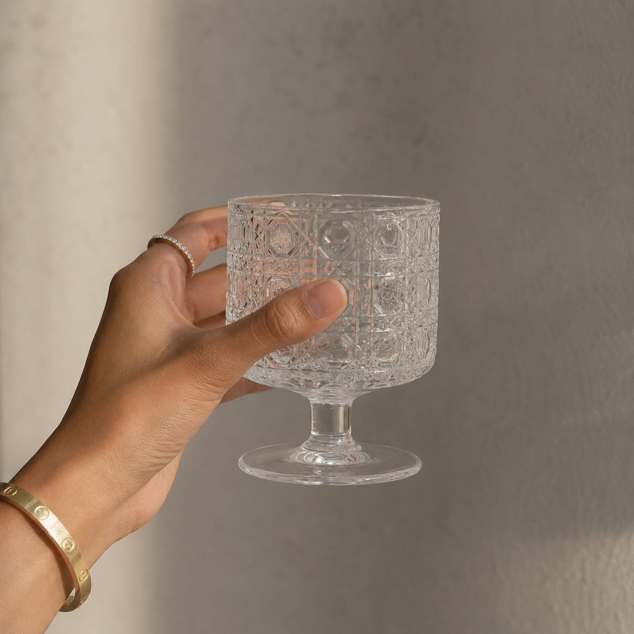 Hand holding a clear glass with intricate patterns against a neutral background