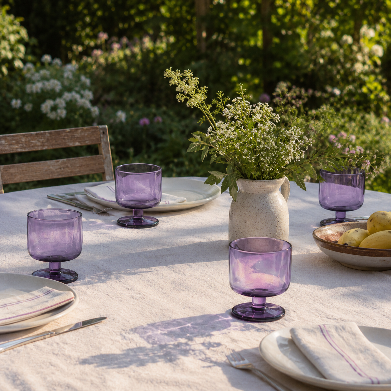 Outdoor dining table setting with purple glasses and a vase of flowers.