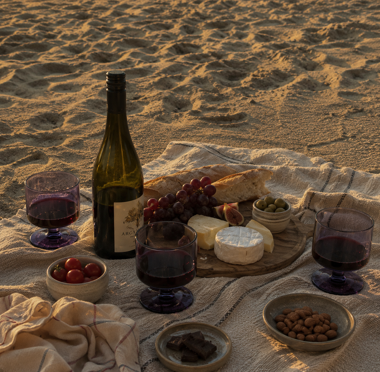 Picnic setup with wine, cheese, and fruits on a sandy beach.