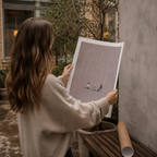 Woman holding a print of a couple sitting on a bench in the rain, standing against a wall outdoors.