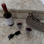 Wine bottle, two glasses with red wine, sunglasses, and a patterned handbag on a textured surface.