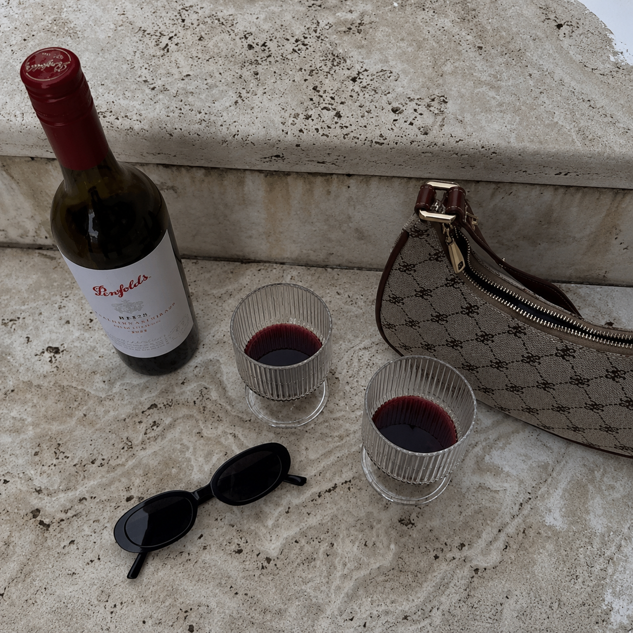 Wine bottle, two glasses with red wine, sunglasses, and a patterned handbag on a textured surface.