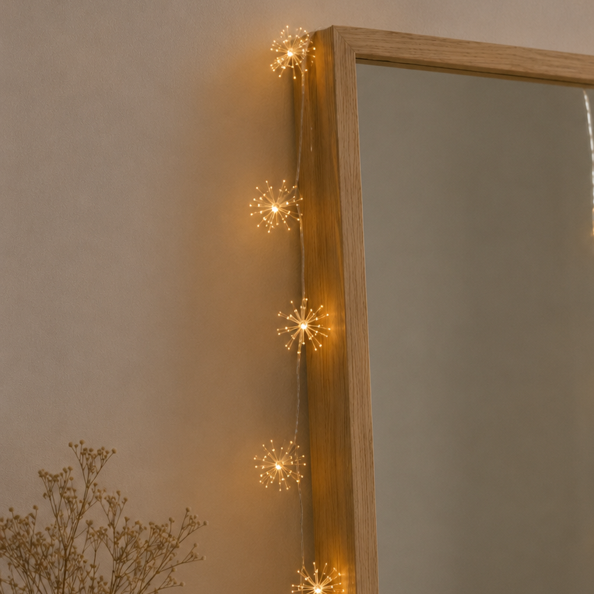 String lights shaped like snowflakes hanging above a mirror with a vase of dried flowers on a neutral wall.
