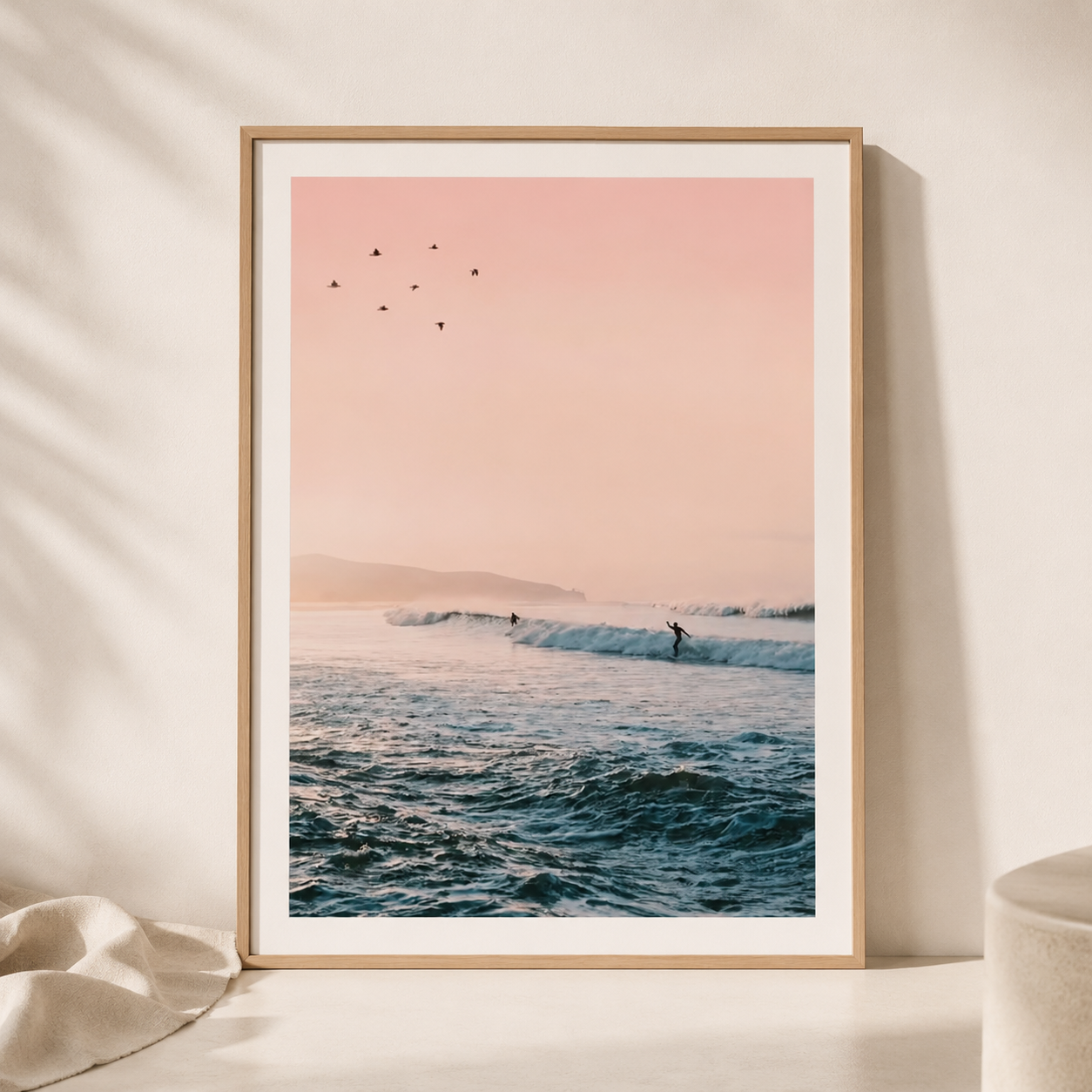 Framed artwork of a beach scene with a pink sky and ocean waves on a beige wall.