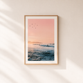 Framed artwork of a sunset over water with birds on a light wall.