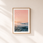 Framed artwork of a sunset over water with birds on a light wall.
