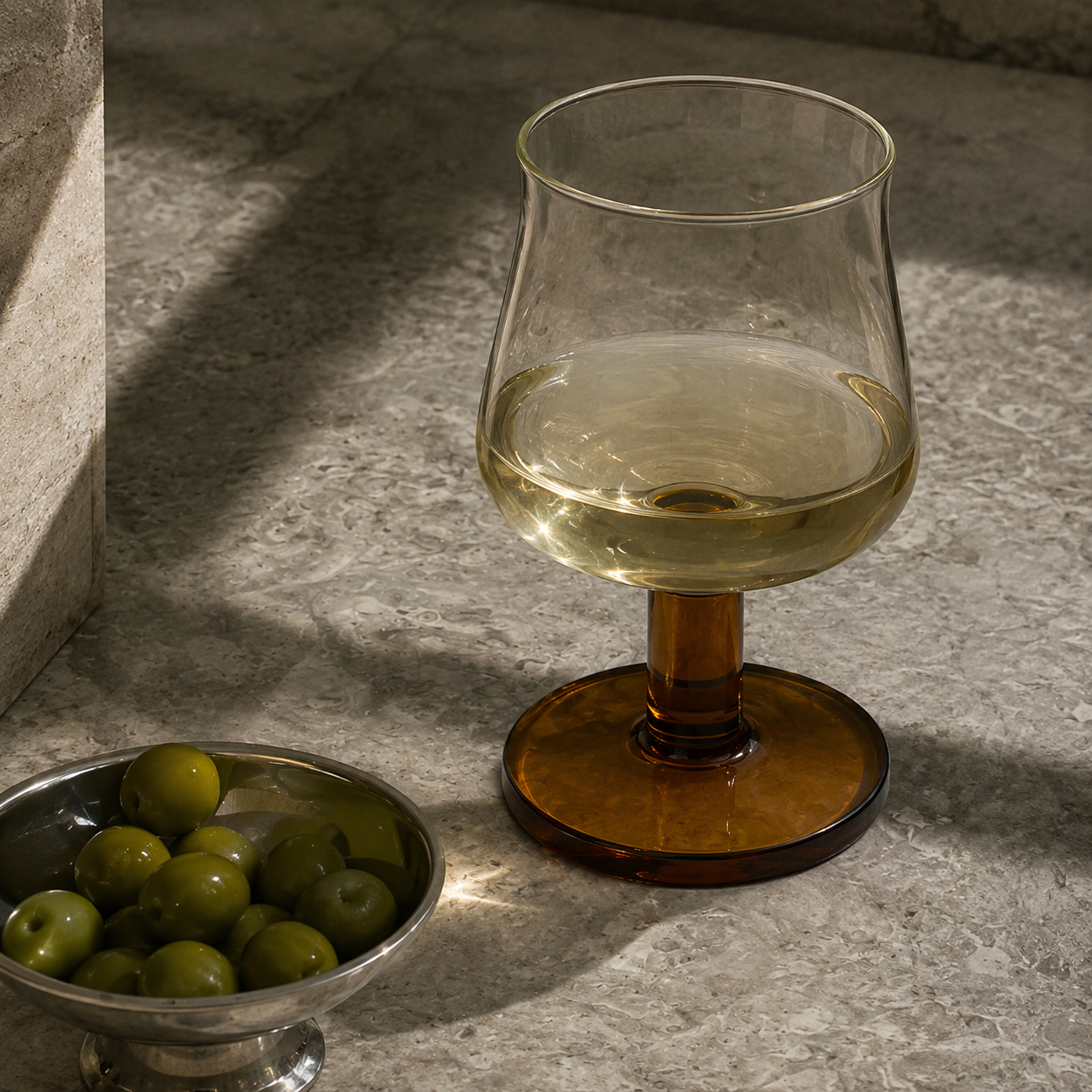Glass with amber base and clear glass filled with a light liquid, next to a bowl of green olives on a stone surface.
