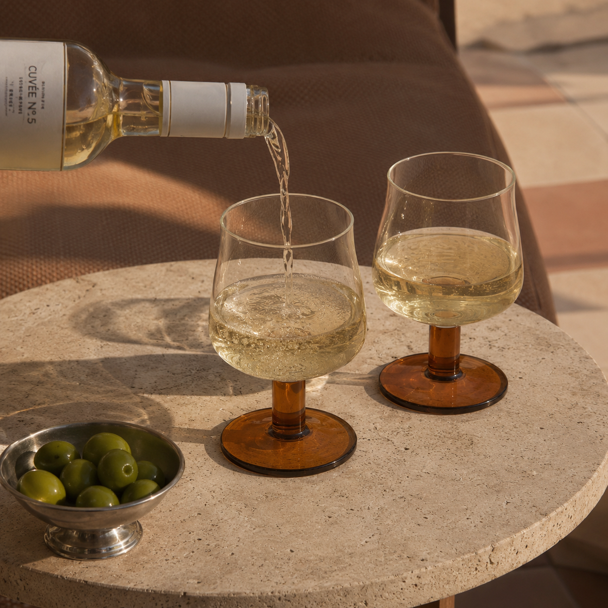 White wine being poured into two glasses on a stone table with a bowl of olives.